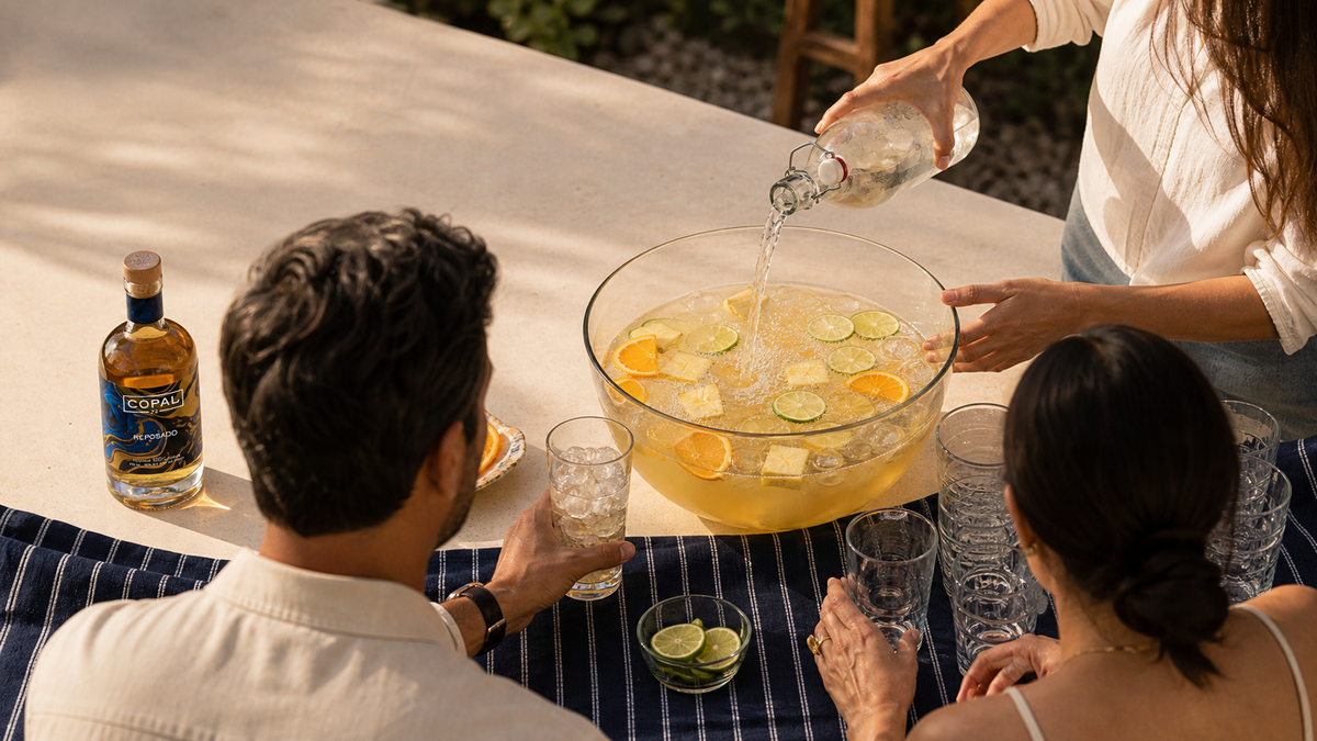 Three adults serving a bright tequila fruit punch from a clear bowl beside a real Copal 22 Reposado bottle at a late-summer backyard table