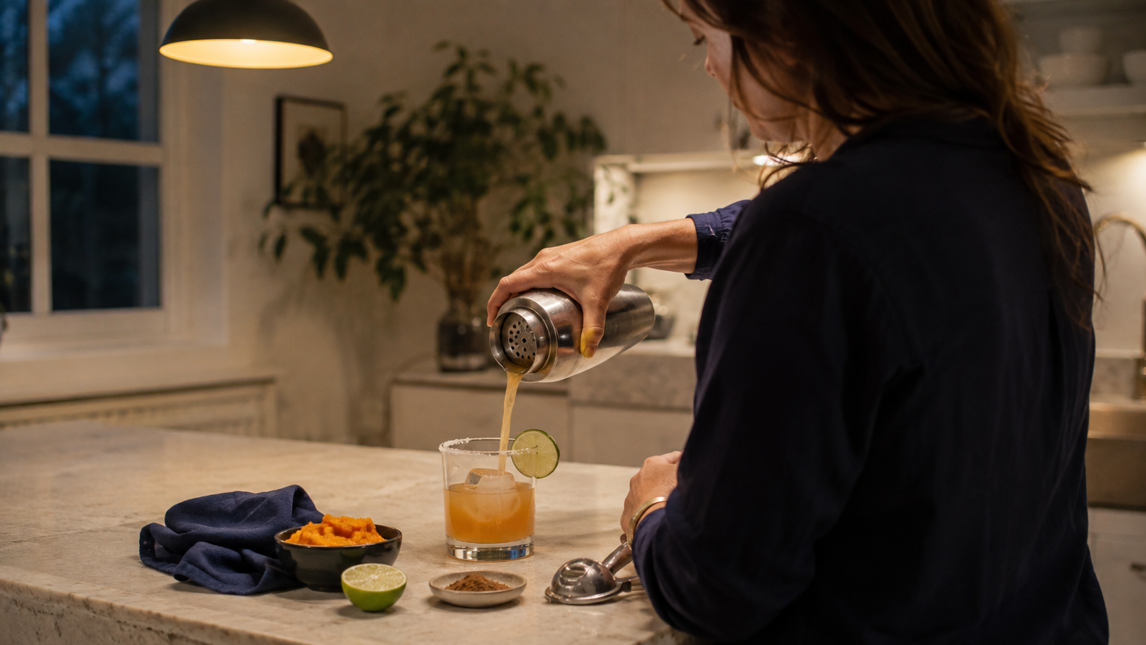 Adult straining a dry pumpkin margarita into a half-salted rocks glass in a warm home kitchen