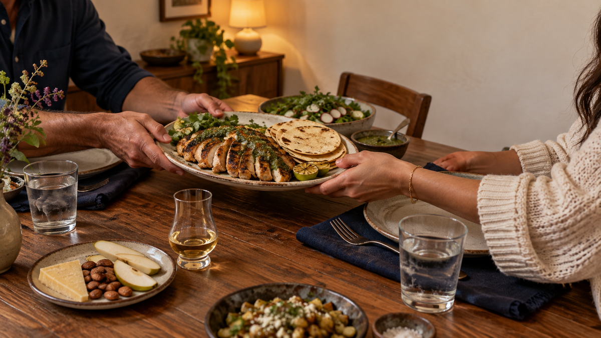 Two adults passing grilled chicken with salsa verde at a warm home dinner beside reposado, Manchego, almonds, and pear